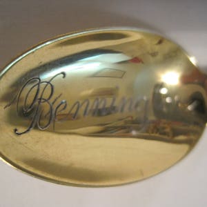 May include: A gold-colored spoon with the engraved name "Benning" on the bowl. The spoon's surface reflects the surrounding environment. The handle is partially visible, featuring a decorative design.