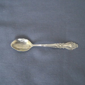 Puede incluir: Una cuchara plateada con un mango ornamentado y un cuenco detallado. El cuenco presenta una imagen grabada de un edificio. La cuchara está sobre un fondo gris oscuro.