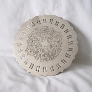 May include: A silver-coloured, round trivet with an ornate, engraved floral design in the centre. The trivet has a scalloped edge with evenly spaced cut-out designs. The background is a white fabric.