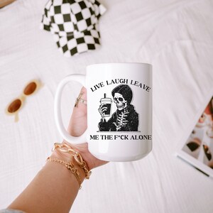 May include: White ceramic coffee mug with a black and white illustration of a skeleton holding a coffee cup. The text on the mug reads "Live Laugh Leave Me The F*ck Alone".
