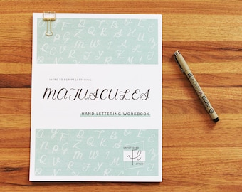 May include: A workbook titled "Majuscules: Intro to Script Lettering" with a light blue background and white lettering. The workbook is open to a page with a grid of uppercase letters in a script font. The page also includes the text "Hand Lettering Workbook" and a logo for "Vaplettered Letters".