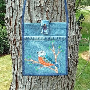 May include: A blue denim crossbody bag featuring a rectangular shape, a single shoulder strap, and a top flap secured with a dark button. The front displays an embroidered design of a bird perched on a branch with small green leaves.