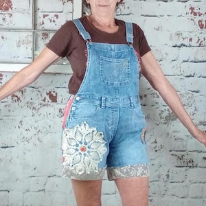 May include: A brown short-sleeve t-shirt paired with light blue denim overall shorts. The overalls feature a large white circular crochet doily appliqué on the right thigh and a decorative lace trim along the bottom hem of the shorts.
