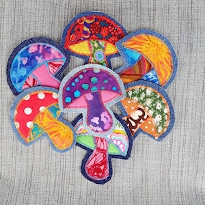 May include: A collection of seven fabric mushroom-shaped appliques, each featuring distinct colorful patterns and textures. The mushrooms are outlined with dark blue denim-like fabric edges and arranged in a cluster on a textured gray surface.