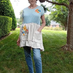 May include: A short-sleeved tunic featuring a light blue upper section and a cream-colored lower section with a ruffled hem. The garment includes a floral applique on the chest and a larger floral embroidery on the side, plus a patch pocket.