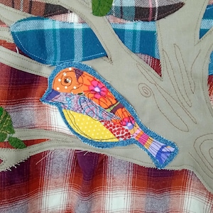 May include: A colorful fabric applique of a bird perched on a tree branch. The bird features orange, yellow, and purple patterned wings and a blue denim outline. The background is a plaid pattern in red, brown, and white.
