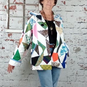 May include: A long-sleeved hooded jacket constructed from a patchwork of various fabric scraps in white, pink, green, orange, blue, and brown. The jacket features a front zipper closure, two side pockets, and a white base with geometric patterns.
