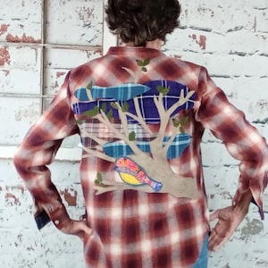 May include: A red and white plaid flannel shirt with a colorful bird and tree design on the back. The bird is perched on a branch with blue and purple accents, and green leaves.