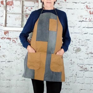 May include: A color-blocked apron featuring vertical panels of tan and charcoal gray fabric. The apron includes two large lower patch pockets and one smaller chest pocket, all constructed from the same tan and gray material with dark straps.