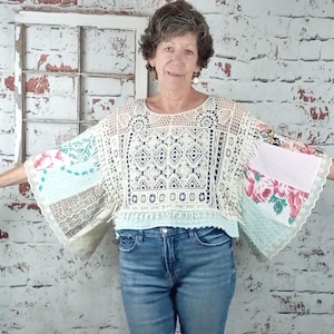 May include: A cream-colored crochet top with patchwork sleeves featuring floral and patterned fabrics. The top has a round neckline and a loose, flowing fit. The model is wearing blue jeans.
