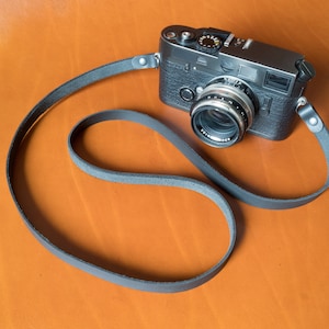May include: A black camera with a silver lens and a black leather strap. The camera is lying on a wooden surface.