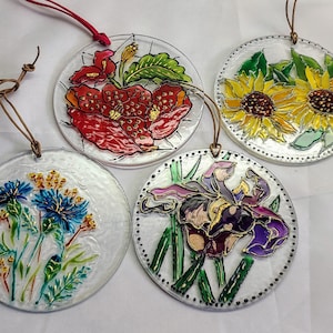 May include: Four clear, circular ornaments with floral designs. One shows a red poppy, another sunflowers, a third blue cornflowers, and the last an iris. Each has a leather cord for hanging.