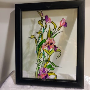 May include: A framed artwork depicting iris flowers. The painting features purple and pink irises with gold outlines and green leaves. The artwork is set against a white background and framed in a black frame, creating a striking contrast.