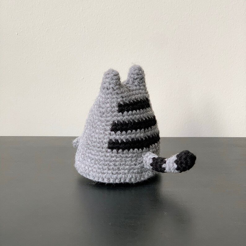 Puede incluir: Un juguete de gato de crochet gris con rayas negras y cola. El gato est&aacute; sentado sobre una superficie negra.