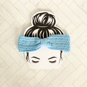 May include: A light blue crocheted headband with a bow. The headband is on a white background with a black silhouette of a woman with a bun.