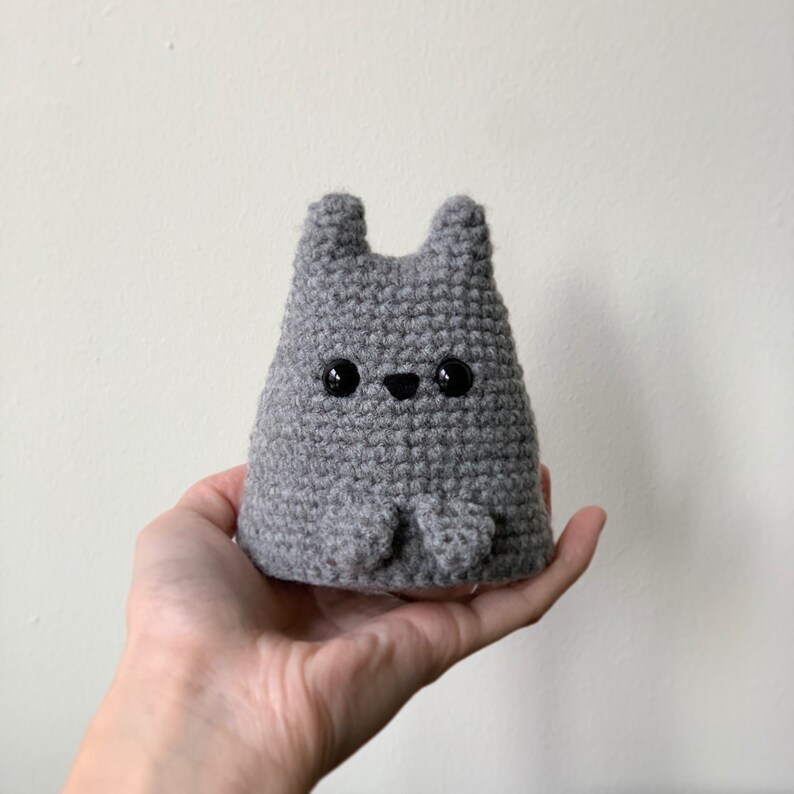 Puede incluir: Un animal de peluche gris de crochet con forma de conejo con ojos negros. El conejo est&aacute; sentado en una mano.