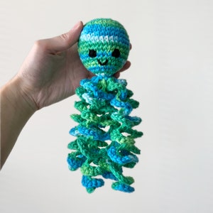 May include: A crocheted octopus toy with a green, blue, and white striped head and a smiling face. The octopus's tentacles are curled and are a mix of green and blue.