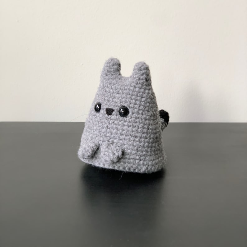 Puede incluir: Peluche de gato gris de crochet con ojos negros y una peque&ntilde;a cola negra.