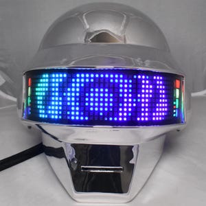 May include: A chrome-finished helmet featuring a curved front visor with a bright blue LED dot matrix display. The display shows a digital pattern of light, with vertical bars of red, green, and blue lights visible on the far left and right edges.