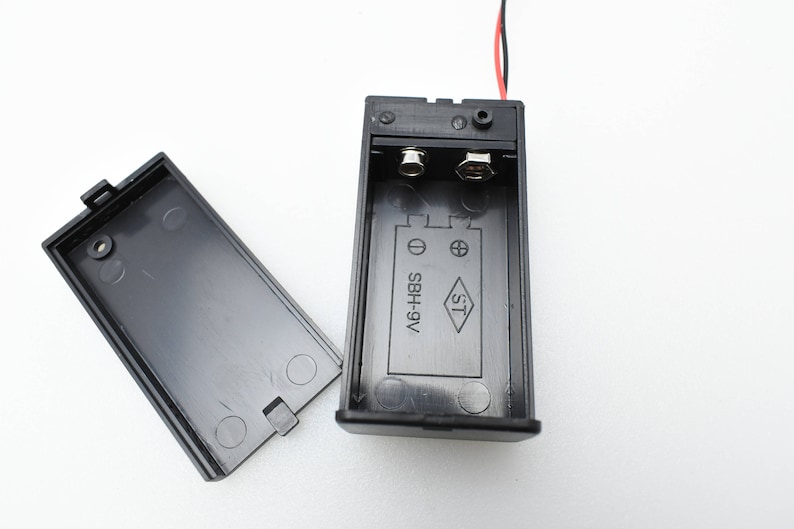 May include: A black battery holder with two red wires attached. The holder has a label that reads "SBH+9V ST".