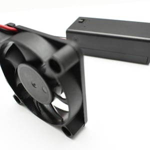 May include: A black computer fan with a black power supply unit. The fan has a black plastic housing and a black blade. The power supply unit has a black plastic housing and a black switch.