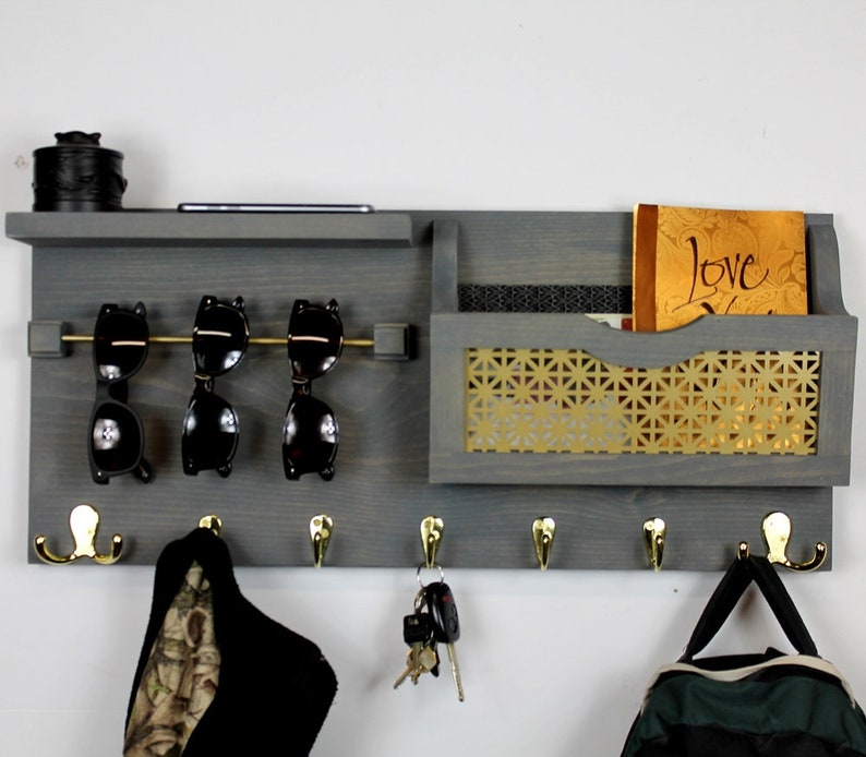 May include: Gray wooden wall organizer with a shelf, hooks, a mail slot, and a gold wire mesh insert. The organizer has a small black box on the shelf, a pair of sunglasses on the hooks, and a book in the mail slot. The book has the word "Love" on the cover.