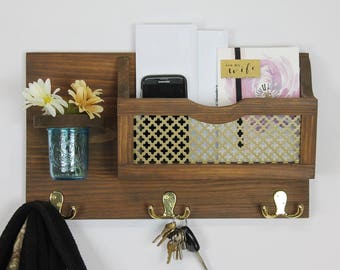 May include: A wooden wall organizer with a key hook, a mail slot, and a small shelf. The mail slot has a gold-colored metal mesh insert. The organizer has a brown finish and is decorated with a small vase of white flowers.