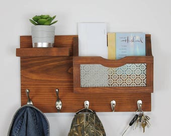 May include: A wooden wall mounted organizer with a shelf, a mail slot, and hooks. The mail slot has a decorative metal front. The organizer is stained a dark brown color. There are three hooks on the bottom of the organizer. A blue jean hat, a camouflage hat, and a set of keys are hanging on the hooks.