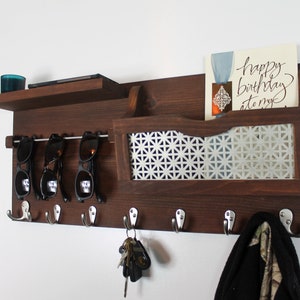 May include: A dark brown wooden wall mounted organizer with a shelf, a mail slot, and hooks. The mail slot has a white patterned metal insert. There are four hooks with keys and a black scarf hanging on them. There are sunglasses on a metal rod above the hooks. The text "happy birthday to my" is visible on a card in the mail slot.