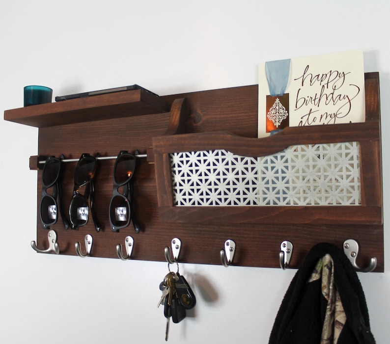May include: A dark brown wooden wall mounted organizer with a shelf, a mail slot, and hooks. The mail slot has a white patterned metal insert. There are four hooks with keys and a black scarf hanging on them. There are sunglasses on a metal rod above the hooks. The text "happy birthday to my" is visible on a card in the mail slot.