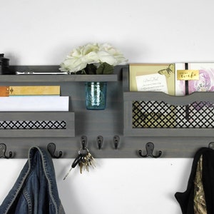 May include: Gray wooden wall organizer with a shelf, two mail slots, and a row of hooks. The mail slots have a black wire mesh insert. The organizer has a small jar with a blue and white pattern on the shelf. There are keys on the hooks and a jacket hanging on one of the hooks. The text "For my wife" is visible on one of the cards in the mail slot.