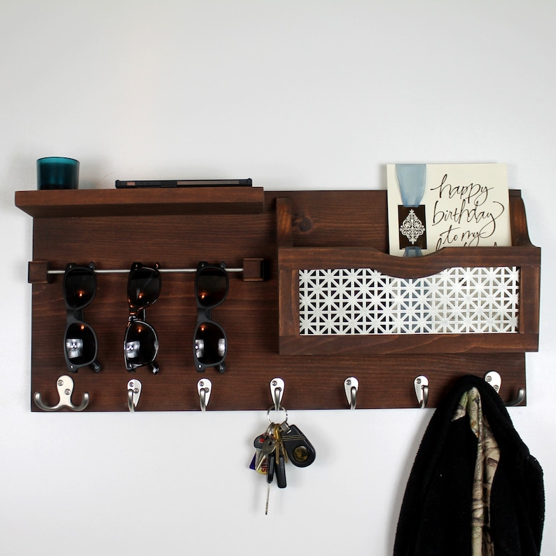 May include: A dark brown wooden wall mounted organizer with a shelf, a mail slot, hooks, and a sunglasses holder. The mail slot has a white patterned metal insert. The organizer has a card with the text "Happy Birthday to my..." in it.