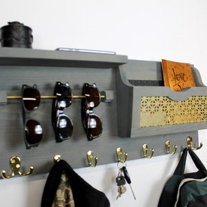 May include: Gray wooden wall organizer with a shelf, a mail slot, a sunglasses rack, and five hooks. The mail slot has a gold mesh backing and the hooks are gold. The organizer has a small black box on the shelf and a book with the word "Love" written on it in the mail slot.