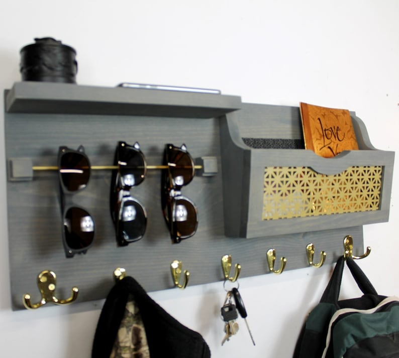 May include: Gray wooden wall organizer with a shelf, a mail slot, a sunglasses rack, and five hooks. The mail slot has a gold mesh backing and the hooks are gold. The organizer has a small black box on the shelf and a book with the word "Love" written on it in the mail slot.
