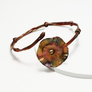 Puede incluir: Un brazalete de alambre de cobre con un gran dije de flor de cobre multicolor. La flor tiene un diseño texturizado y en capas con un pequeño punto dorado en el centro.
