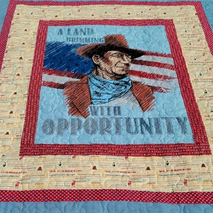 May include: A quilted wall hanging featuring a portrait of a cowboy with the American flag. The text reads "A Land Brimming With Opportunity." The quilt has a blue background and a red border with repeating text and star patterns.