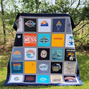 May include: A handmade quilt made from repurposed t-shirts. The quilt features a variety of colorful squares with logos and text, including "Pensacola Mess Hall", "Lifeguard", and "Auburn". The quilt has a gray border and a dark blue binding.