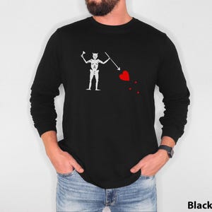Puede incluir: Una camiseta negra de manga larga con un gráfico blanco de calavera y huesos de pirata. El gráfico presenta un esqueleto sosteniendo una lanza y un corazón rojo con tres puntos.
