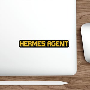 May include: A rectangular sticker with the words "HERMES AGENT" in yellow, pixelated font, outlined in black. The sticker is on a white surface, with a wooden desk and a computer mouse visible in the background.