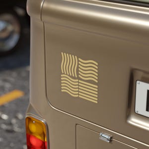 May include: A beige decal featuring a square design composed of four quadrants of wavy lines, applied to the metallic surface of a vehicle. The graphic consists of vertical and horizontal wave patterns in a light tan color against the beige body.