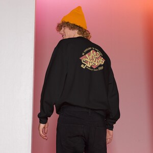 May include: Black long-sleeve shirt with a red and yellow graphic on the back that says "Wild Stallions World Tour 1989". The shirt is being worn by a person wearing a yellow beanie.