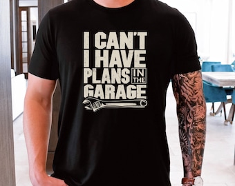 May include: Black t-shirt with the text "I CAN'T I HAVE PLANS IN THE GARAGE" in white block letters. A white wrench graphic is below the text. The shirt is a crew neck style.