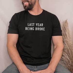 Könnte beinhalten: Schwarzes T-Shirt mit Rundhalsausschnitt und dem weißen Text "LAST YEAR BEING BROKE". Das Shirt besteht aus einem weichen Material und eignet sich für Freizeitkleidung. Das Model trägt eine blaue Jeans.