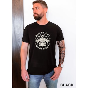 Könnte beinhalten: Ein schwarzes T-Shirt mit einem weißen Grafikdesign. Die Grafik zeigt eine Person in einer Kapuzenrobe mit Würfeln und dem Text "Dad by Day DM by Night".