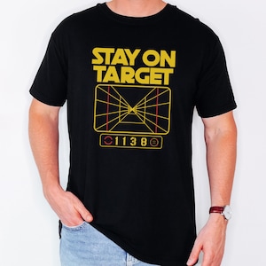 Pode incluir: Camiseta preta com um gráfico amarelo que diz "STAY ON TARGET" e apresenta um sistema de mira do filme Star Wars. O gráfico também inclui os números "011380".