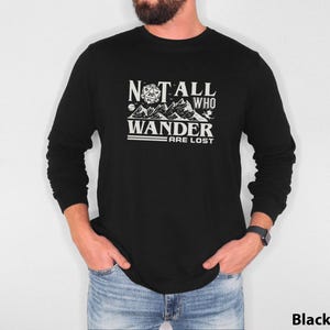May include: Black long-sleeved top featuring the white text "NOT ALL WHO WANDER ARE LOST". The design includes mountains and a 20-sided die. The shirt has a crew neck.