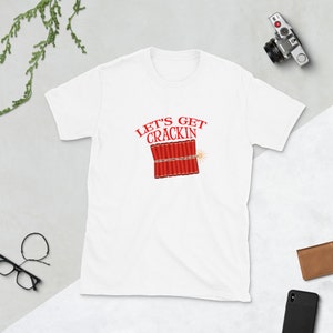 May include: White t-shirt featuring the red text "LET'S GET CRACKIN" above a graphic of red dynamite sticks with a lit fuse. The shirt has a crew neck and short sleeves.