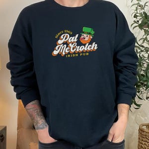 Op de afbeelding: Een zwart sweatshirt met een grafisch ontwerp van een cartoon-kabouter met de tekst "Since 1969 Pat McCrotich Irish Pub".