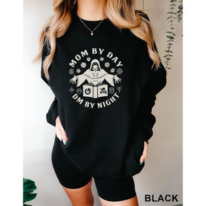 May include: Black sweatshirt with a white graphic design that says "Mom by Day DM by Night" with a stylized image of a person in a hooded robe holding a book and dice.