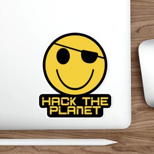 May include: A yellow smiley face sticker with a black outline and a black eye patch. The sticker has the text "HACK THE PLANET" in a bold, futuristic font on a black rectangle below the smiley face.